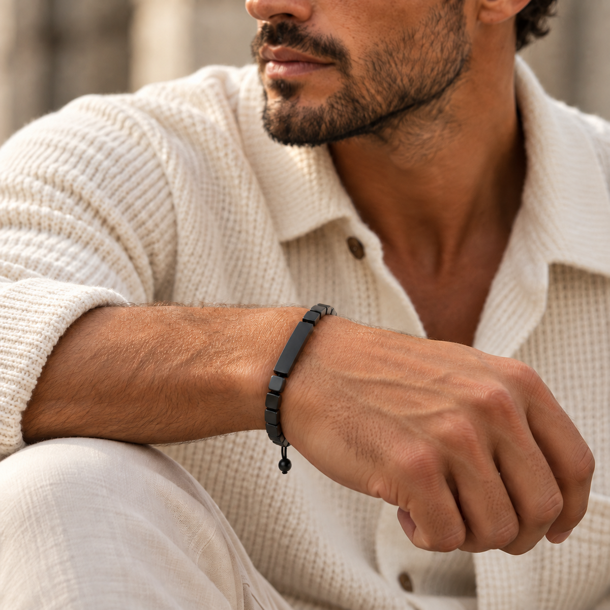 A close-up of a man wearing a black rectangular beaded bracelet on his wrist, dressed in a cream-colored knit shirt.
